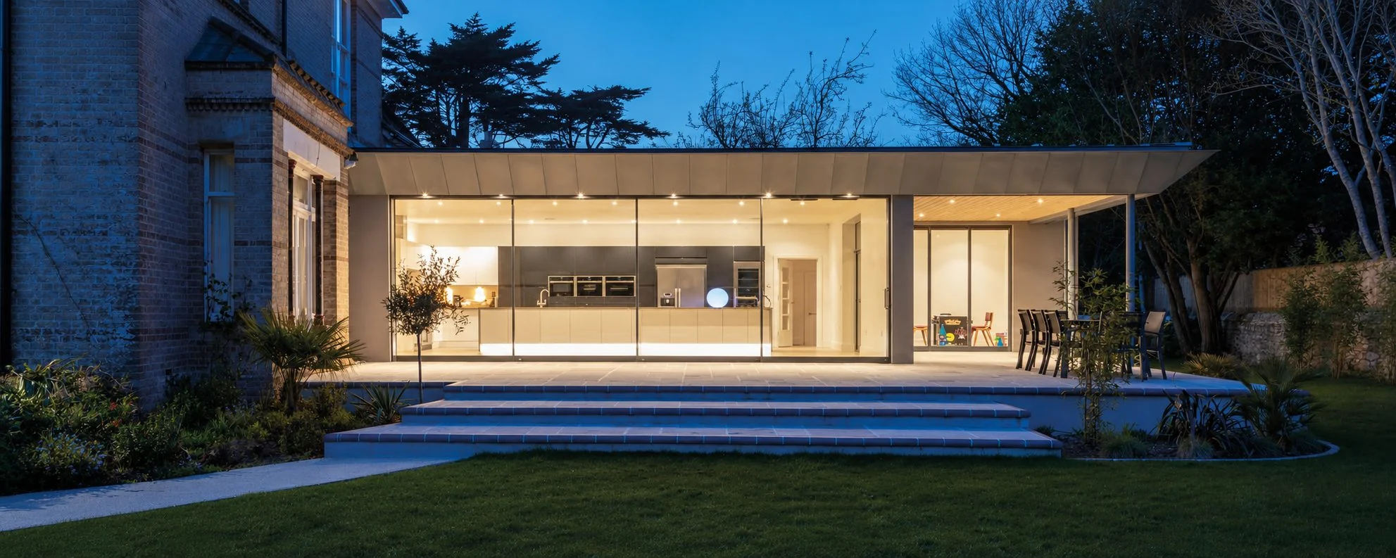 Panoramic view of the stone house and illuminated glass extension opening onto garden steps